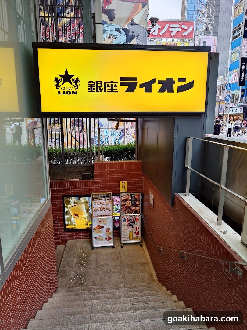 A bright yellow sign with 'GINZA LION' and Japanese characters is mounted above a staircase entrance to a shop. The building facade features red brick walls, metal railings on the right, and glass windows on the left. Below the main sign, there are promotional posters and menu boards displaying food items like burgers and drinks.
