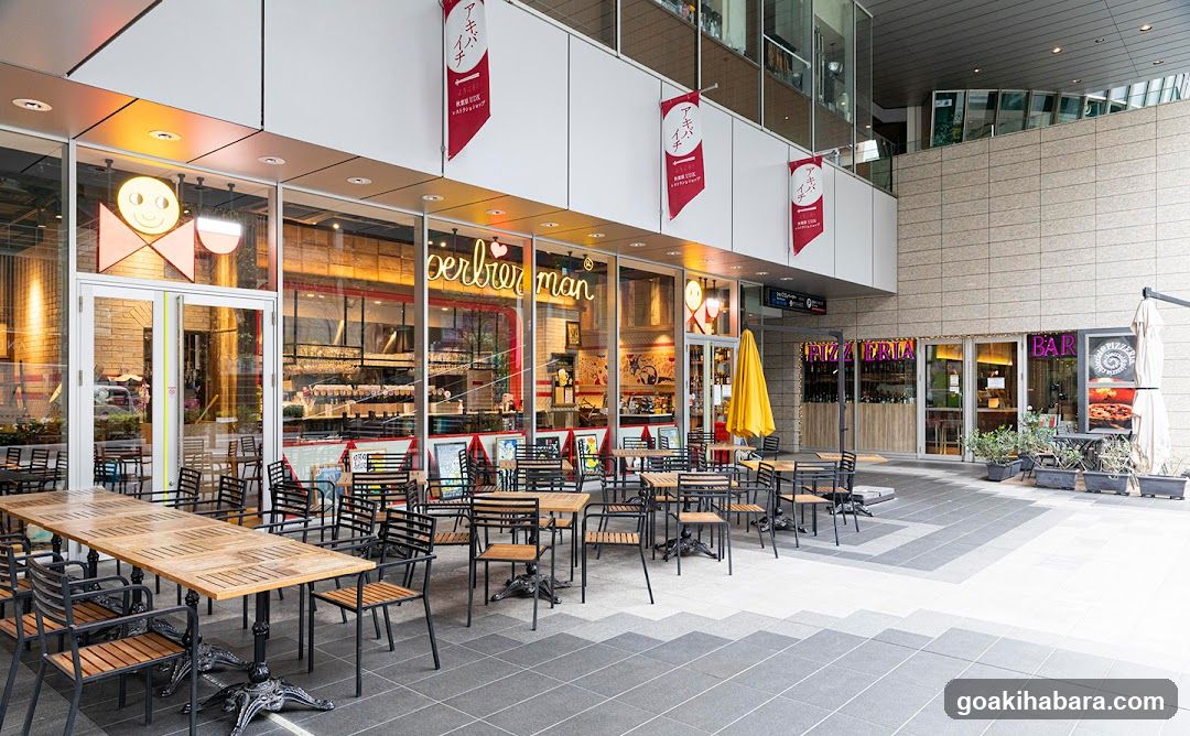 A modern brewery storefront featuring large glass windows displaying interior shelves and equipment. Red banners hang above the entrance, each bearing Japanese characters and a logo. A neon sign reads 'beerbierman' in cursive script across the window. Outside, wooden tables and black metal chairs are arranged on a tiled patio under an open-air structure. To the right, another establishment labeled 'BAR' is visible with purple signage.