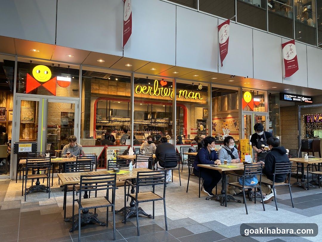 Photo of the physical exterior entrance and storefront facade of Oerbeerman, a bakery/cafe chain. Visible signage includes illuminated 'oerbeer man' neon lettering on the glass front, red banners with Japanese characters hanging above, and yellow smiley face signs flanking the entrance. Large windows reveal interior kitchen equipment and customers seated at outdoor tables. A small sign near the door reads 'Cafe & Bakery'. No menu boards are visible in this shot.