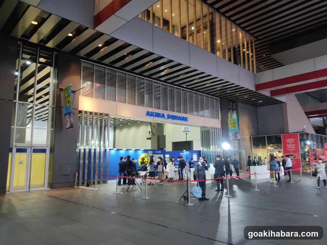 Exterior view of Akiba Square entrance at night, featuring a modern facade with glass panels and metal accents. A blue sign reading 'AKIBA SQUARE' is prominently displayed above the entrance. People are gathered near the entrance, some standing in line behind red ropes. The area is well-lit by artificial lighting.