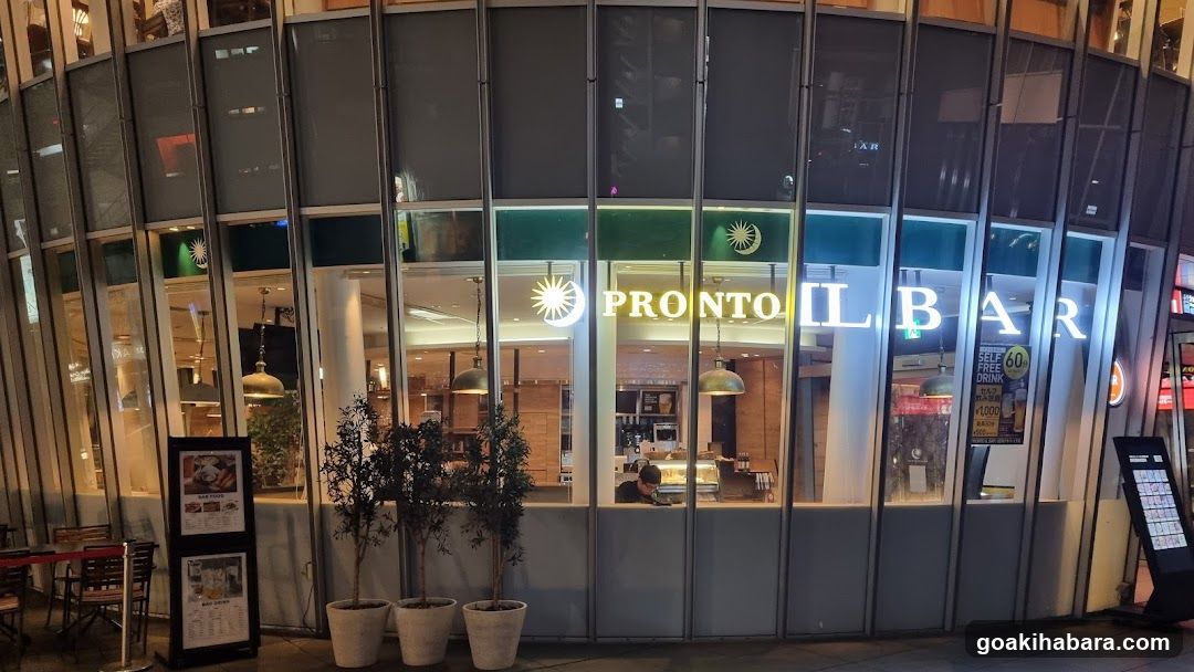 Night view of a modern glass-fronted bar with illuminated signage reading 'PRONTO BAR'. Two potted plants stand outside near the entrance. A digital menu board is visible on the right side, and another framed poster stands to the left.