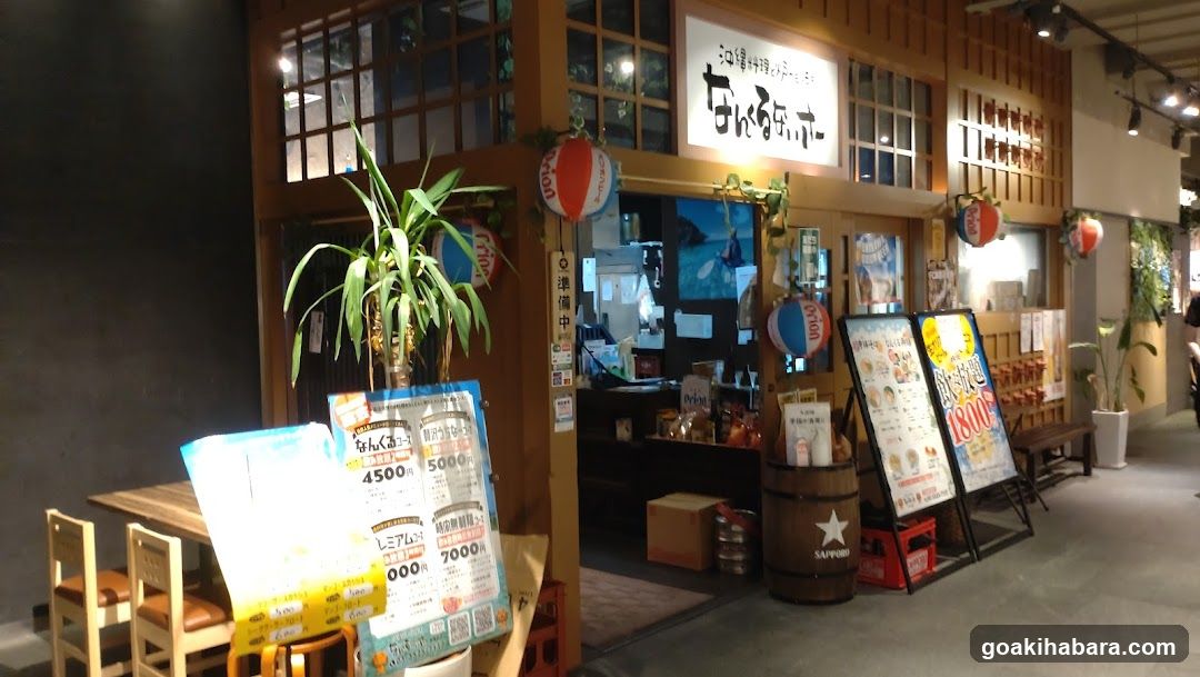 The image shows the exterior entrance of a restaurant or shop, featuring a wooden facade with large windows and a signboard displaying Japanese characters. There are decorative elements such as hanging lanterns and potted plants near the entrance. A menu board is visible outside, listing prices and items.