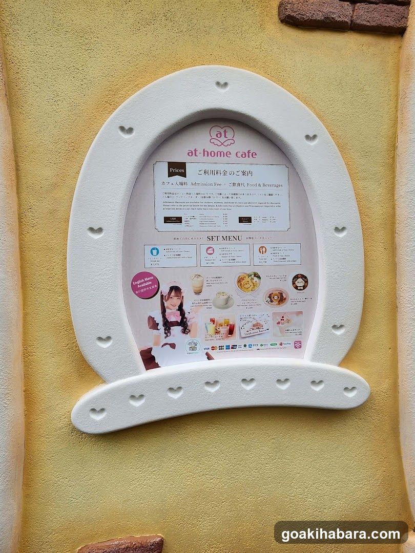 A white, horseshoe-shaped menu board mounted on a yellow stucco wall with heart cutouts along the edges. The board displays 'at-home cafe' branding and includes sections for prices, admission fees (free), set menus with food images, and promotional content featuring a character illustration.