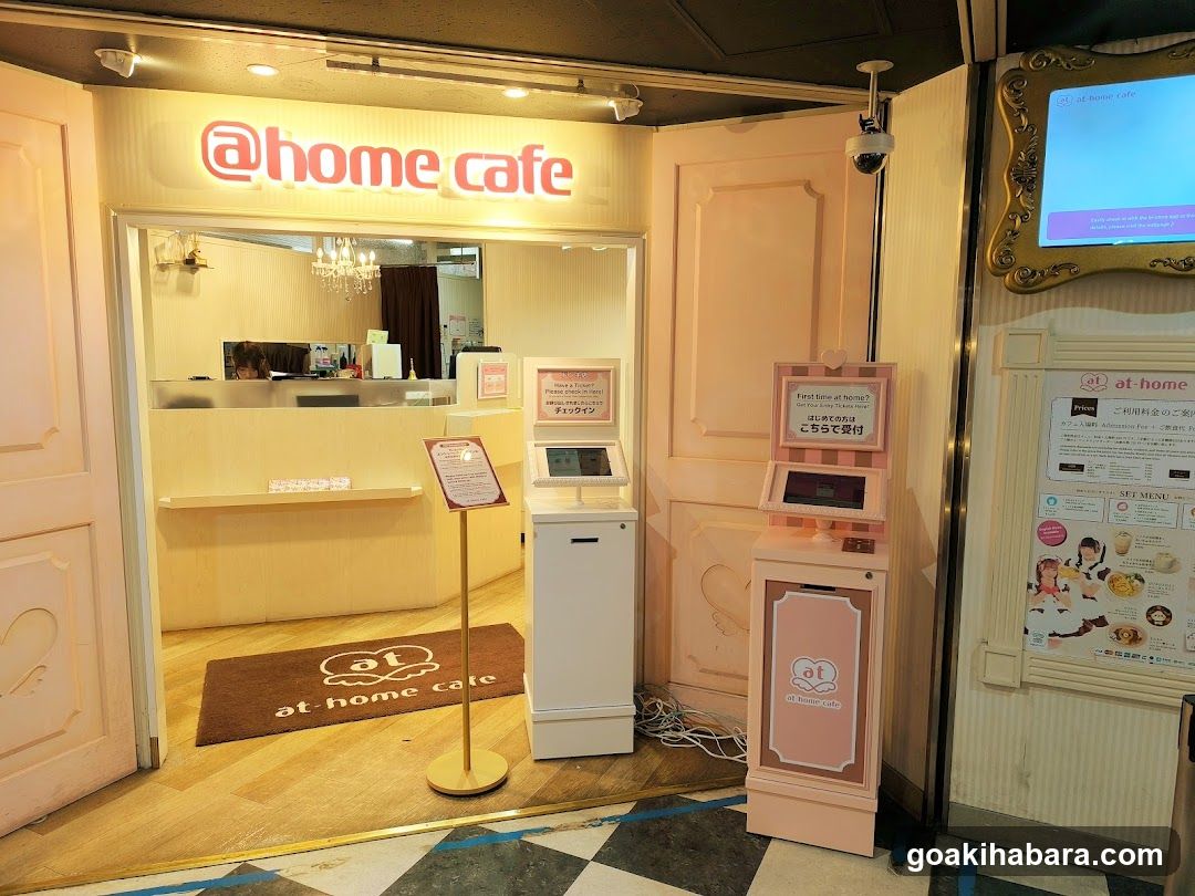 Entrance to @home cafe with illuminated signage above door, pink and cream color scheme, self-checkout kiosk on right, welcome mat at threshold, interior visible through glass showing counter area.