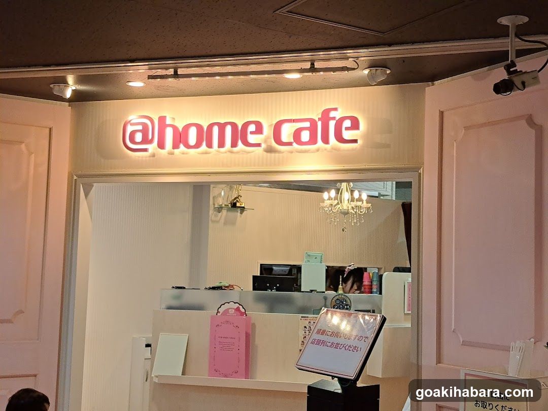 The photo shows the entrance of a shop called '@home cafe'. The sign is illuminated in pink and white, mounted above the doorway. Inside, there is a counter with a computer monitor, a chandelier hanging from the ceiling, and various items on display including a pink menu standee and a small sign with Japanese text.