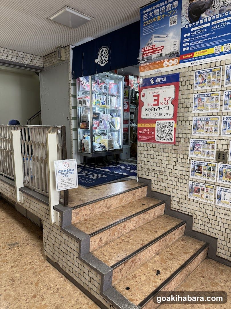 A photo of a convenience store entrance featuring glass display cases filled with products, a blue awning with Japanese characters, and promotional posters advertising PayPay discounts. The wall is tiled with small white tiles, and there are stairs leading up to the entrance. A sign prohibits smoking inside.