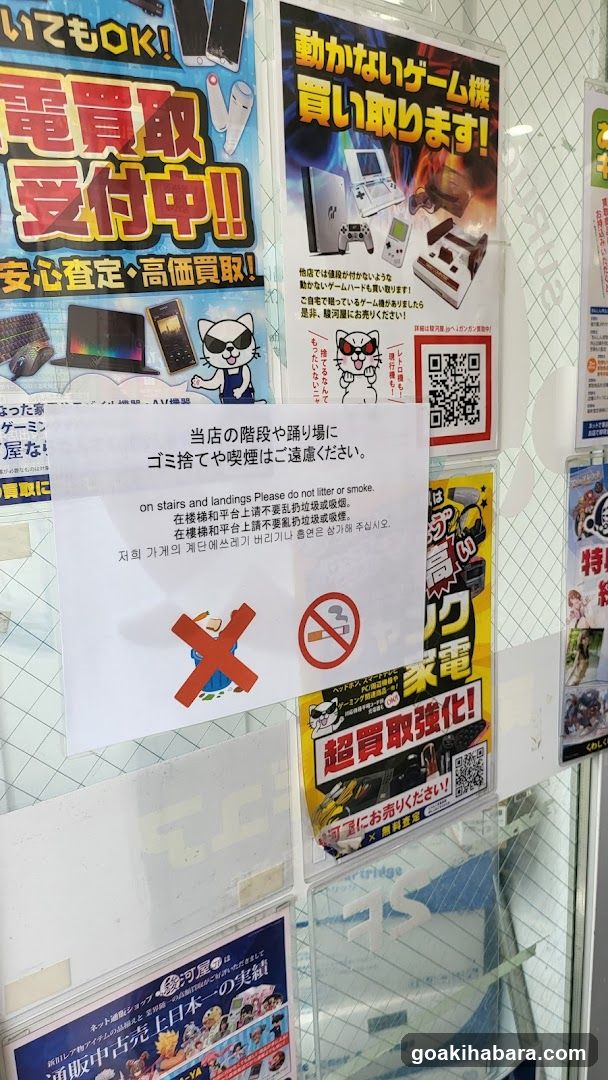 A storefront with multiple posters advertising electronics sales, including game consoles and TVs. A notice prohibits littering and smoking on stairs and landings.
