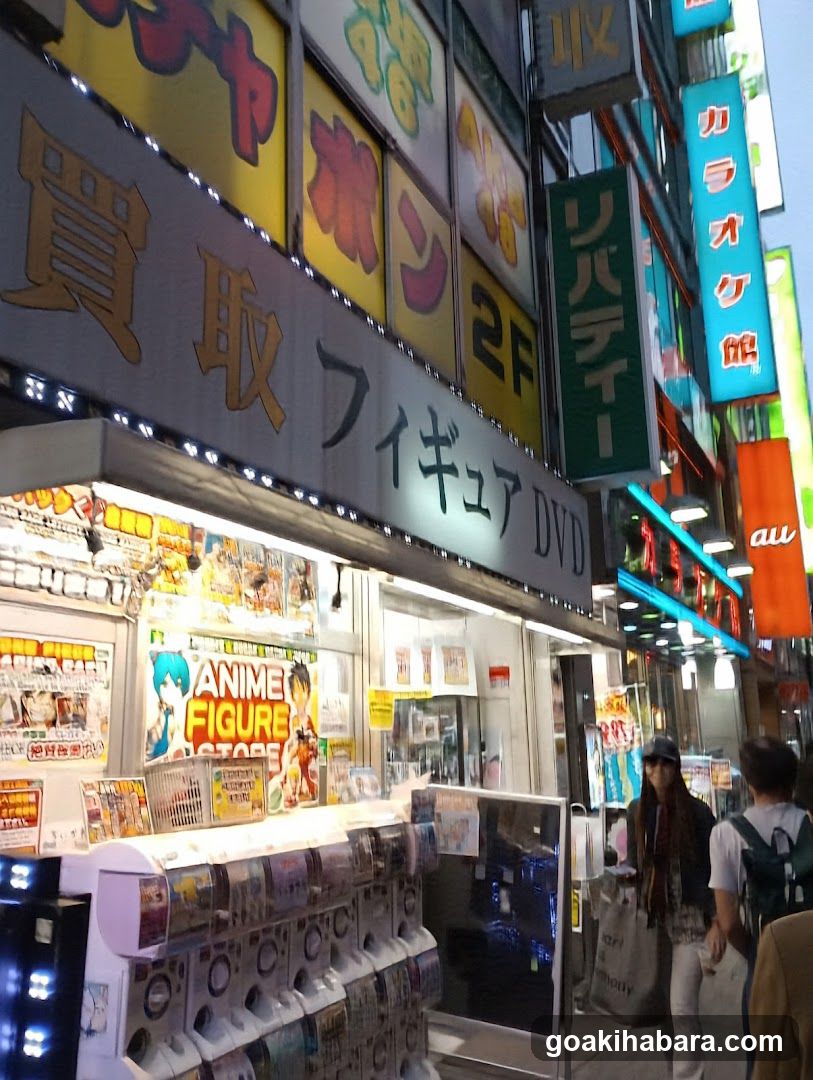 A brightly lit storefront with large Japanese signage reading 'Buy' and 'Figure DVD'. The shop displays anime figures, manga, and merchandise under illuminated signs including '2F', 'Liberty', and 'Karaoke'. A person walks past the entrance.