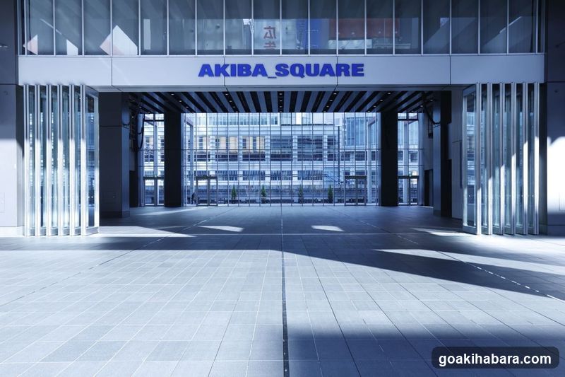 The photo shows the exterior entrance of Akiba Square, a modern building with large glass windows and metal framing. The sign 'AKIBA SQUARE' is prominently displayed above the entrance. The area appears clean and well-maintained, with sunlight casting shadows on the tiled floor.