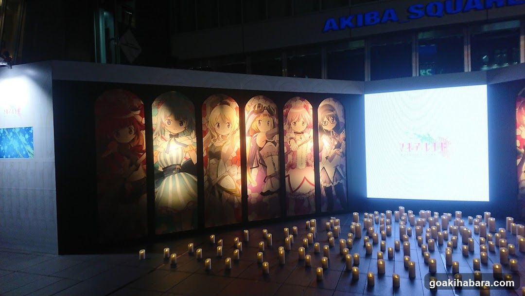 Nighttime exterior view of Akiba Square building with illuminated anime character posters on a curved wall and numerous candles arranged on the ground in front.