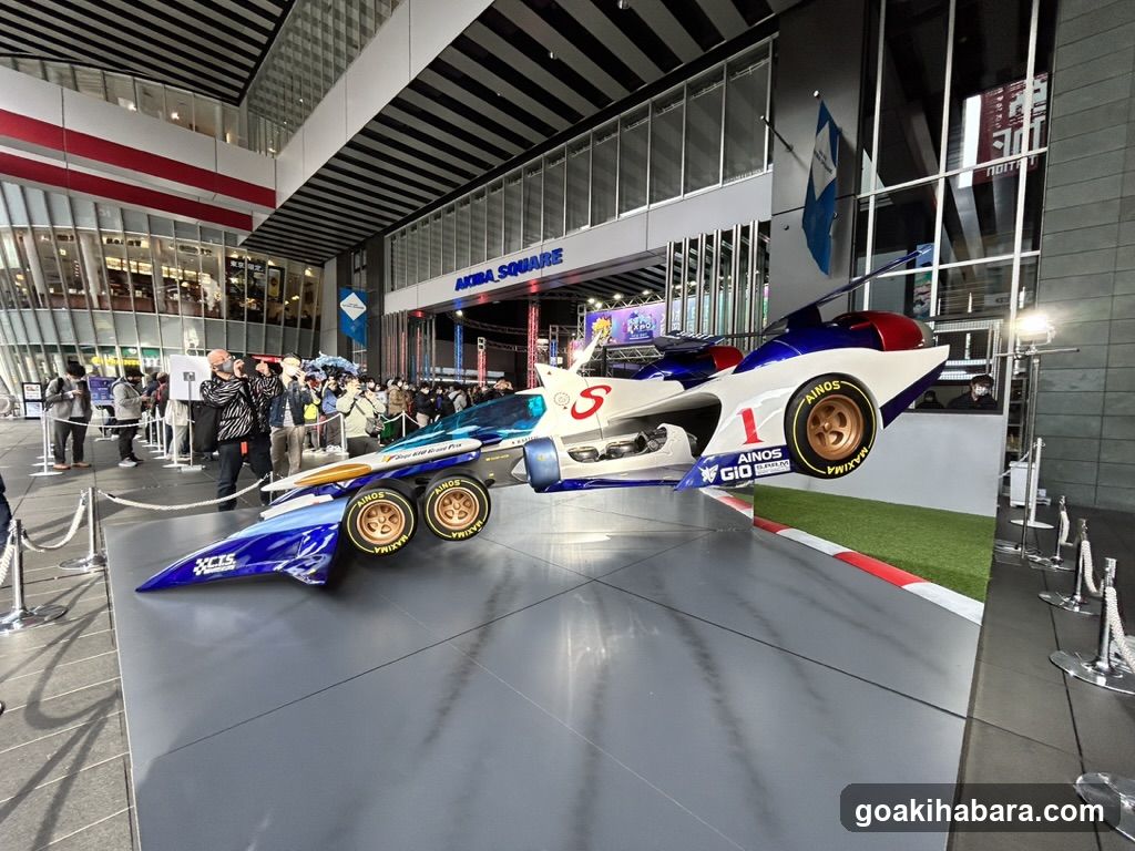 Photo of physical exterior entrance/storefront facade with visible shop signage 'AURORA SQUARE' on building. Features a large display of futuristic racing cars, including blue and white models with sponsor logos like 'ANOS', 'GIO', and 'PRIVAT'. People are gathered around the exhibit inside a modern glass-fronted structure.