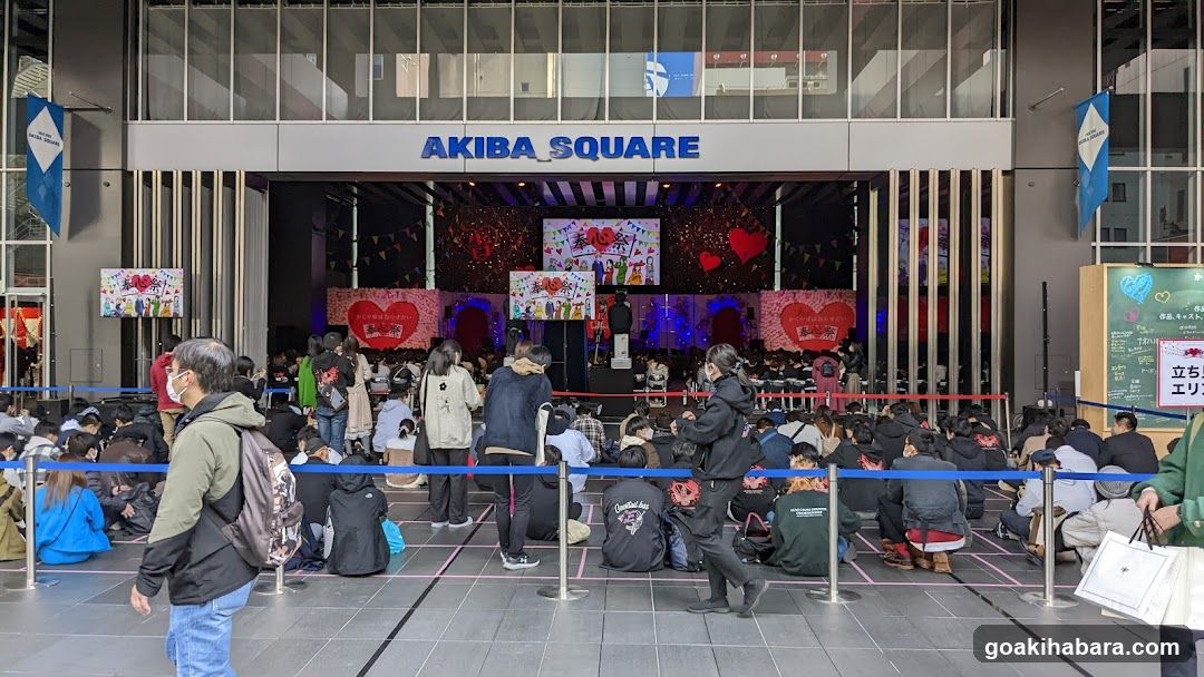 Photo of the physical exterior entrance to Akiba Square, featuring a large stage setup with heart-themed decorations and banners. A crowd is gathered in front, seated on the ground behind blue ropes. Visible signage includes 'AKIBA SQUARE' above the entrance and Japanese text on side panels.