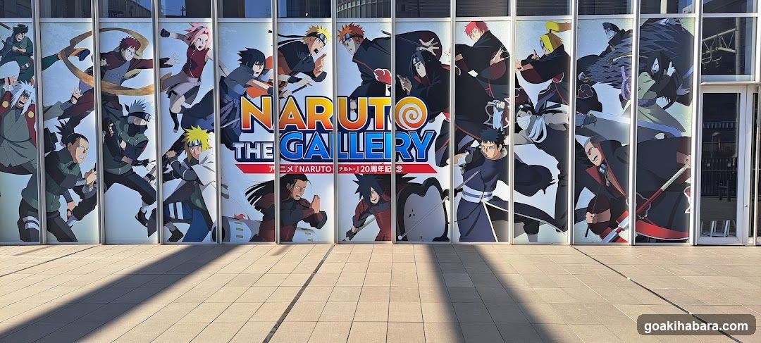 A building facade featuring large glass panels decorated with colorful anime characters from the Naruto series, including Naruto Uzumaki and other ninja figures in dynamic poses. Central signage reads 'NARUTO THE GALLERY' with Japanese text below indicating a 20th anniversary celebration. The entrance is visible on the right side.