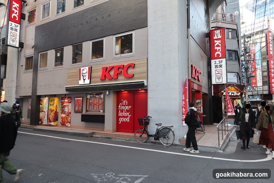 A KFC restaurant exterior with red and white branding, visible signage including 'it's finger lickin' good', a menu board on the wall, posters of food items, and pedestrians walking nearby.