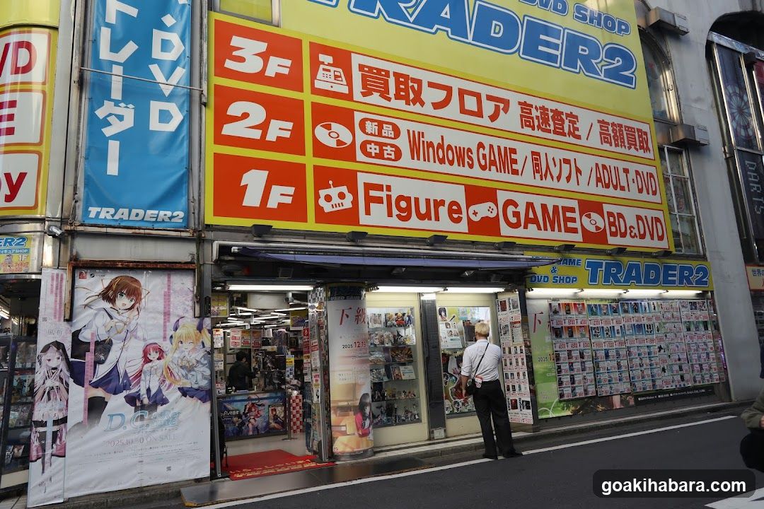 A street-level view of a Japanese video game and anime merchandise store named TRADER2. The facade features large yellow signage with red and white panels indicating different floors (1F, 2F, 3F) selling items like Windows GAMES, ADULT-DVDs, Figures, BD&DVDs. A poster on the left displays anime characters. A man stands near the entrance looking at merchandise inside.