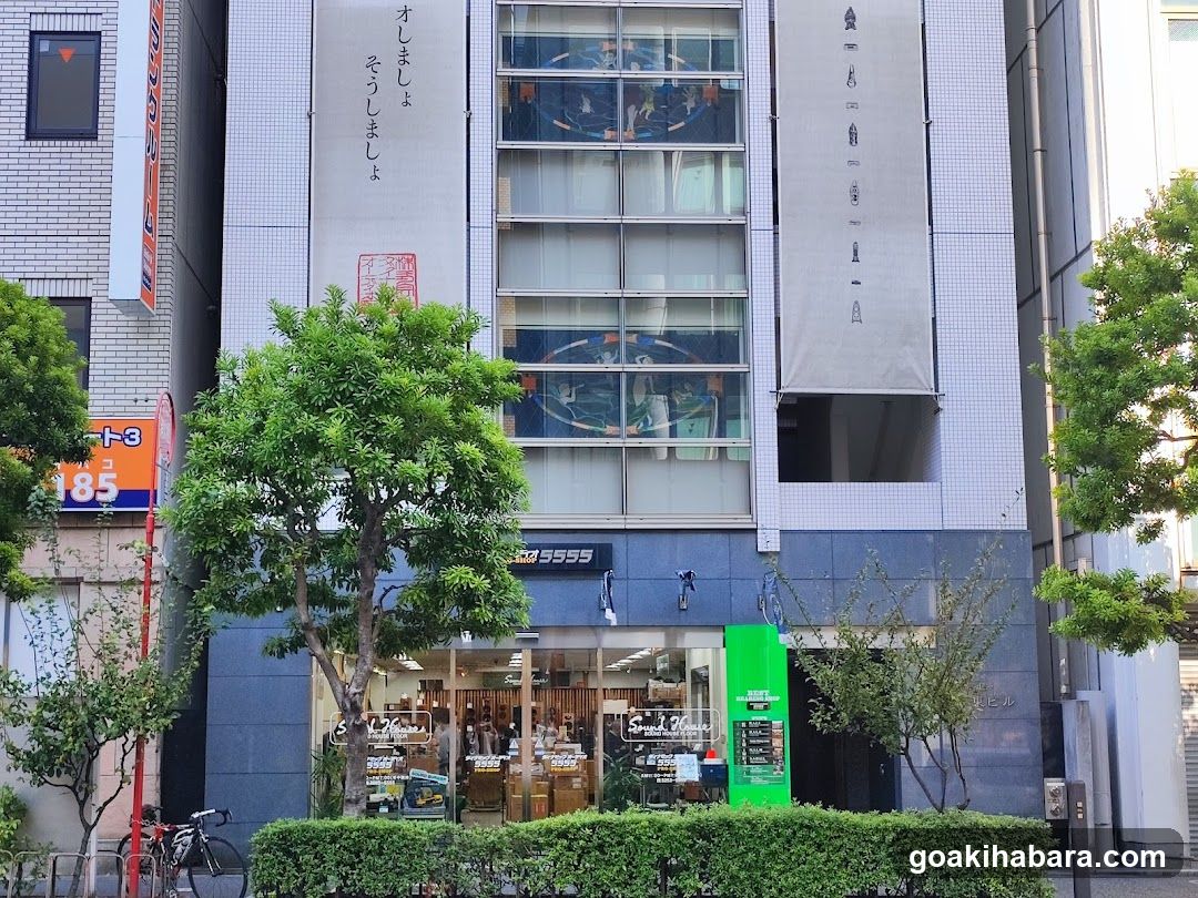 A street-level view of a commercial building featuring a shop entrance with glass doors, wooden interior elements, and visible signage including '5555' above the door. A bright green kiosk stands to the right near the entrance. Trees frame the scene on both sides, and vertical banners with Japanese characters hang from upper windows. The ground floor appears open to pedestrians, with bicycles parked nearby.