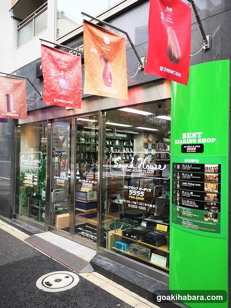 The image shows the exterior of a shop called 'Sound House' with glass doors and windows displaying audio equipment inside. Hanging banners advertise products, including earbuds. A green pillar on the right lists events or promotions.