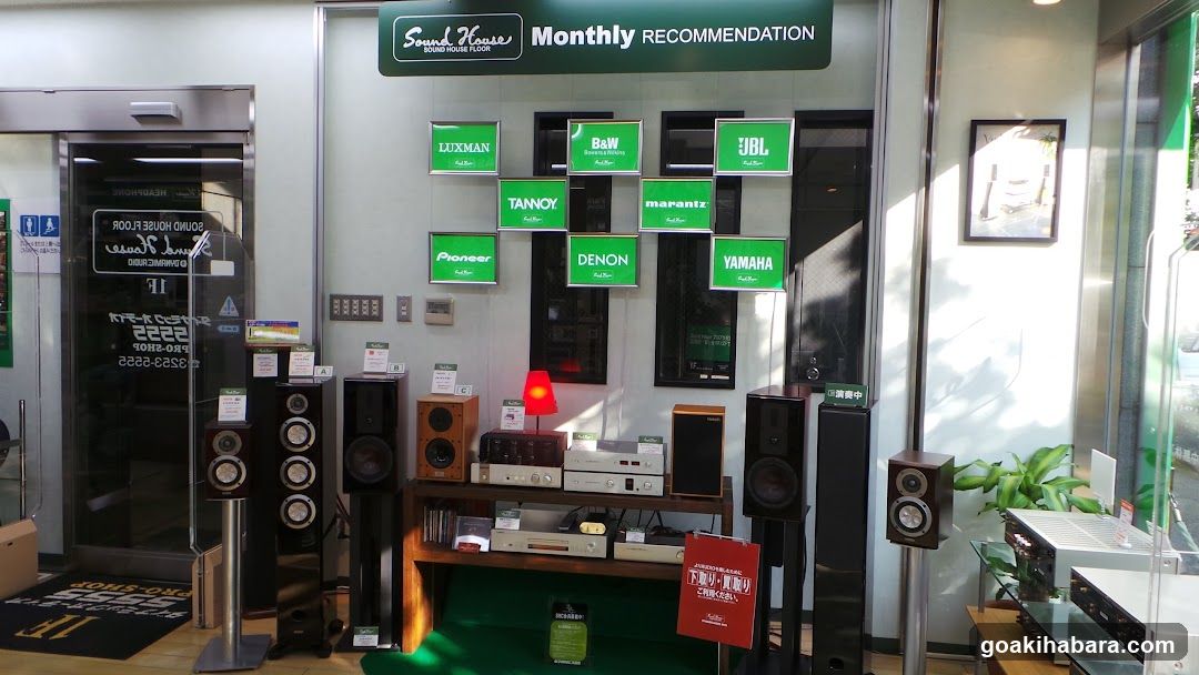 Exterior storefront of a sound equipment shop featuring a display of audio gear including speakers, amplifiers, and receivers under a 'Monthly RECOMMENDATION' sign. Brand logos like JBL, Yamaha, Denon, Marantz, Tannoy, Luxman, Pioneer, Baow are visible on green signs. A glass door entrance with signage is to the left.