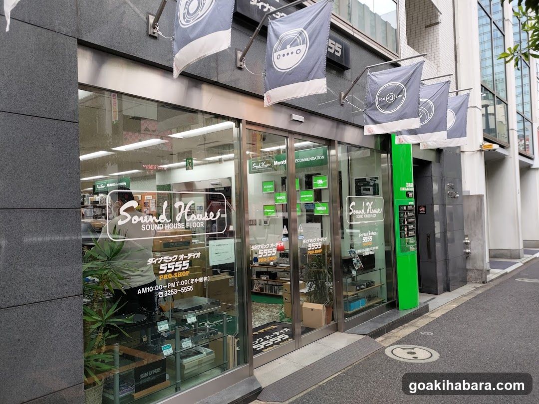 A modern shop front with large glass windows and doors, displaying 'Sound House' signage on the facade. Flags hang above the entrance. Inside, shelves of products are visible through the glass.
