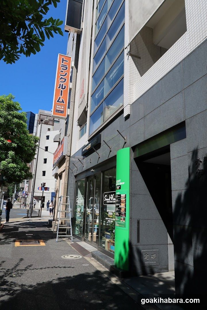 A shop front on a city street, featuring glass doors, a green vertical signboard, and an orange hanging sign with Japanese characters. A ladder is placed near the entrance.