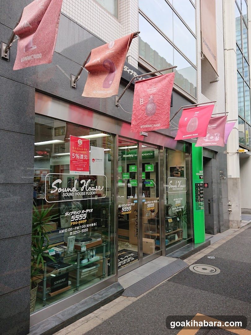 The image shows the exterior of a shop called 'Sound House' with large glass windows and doors. Pink banners hang above, displaying the store's logo and Japanese text. Inside, shelves are visible with products, including what appears to be audio equipment. A sign on the window advertises a 5% discount.