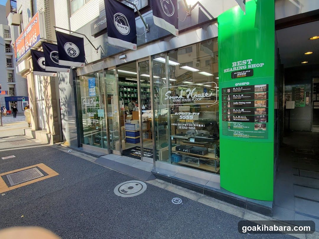 A street-level view of a shop named 'Sound House' with large glass windows and doors, displaying audio equipment inside. Flags hang above the entrance. A green pillar on the right features a menu board listing services like HALLI and audio fitting rooms.