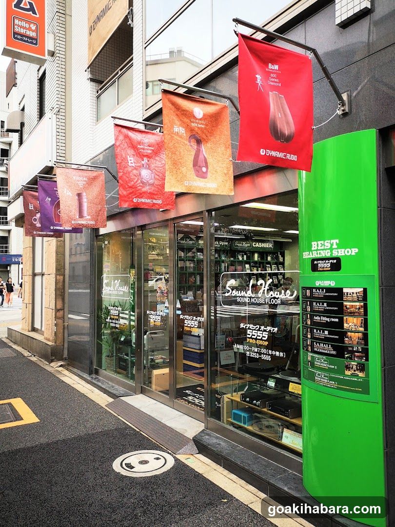 A street-level view of a music store with glass doors and windows displaying audio equipment inside. Hanging banners advertise products like headphones. A green pillar on the right lists sale items with prices in yen.