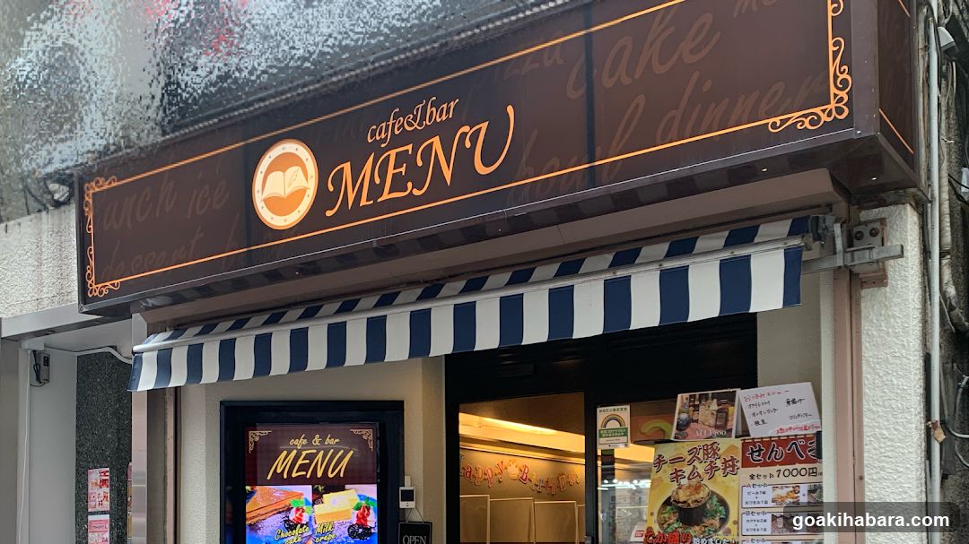 A shop front with a large brown sign displaying 'cafeLbar MENU' and decorative script text like 'cake', 'dinner', 'ice cream'. Below, a blue-and-white striped awning covers the entrance. A smaller menu board is visible in the window, showing food items and prices.