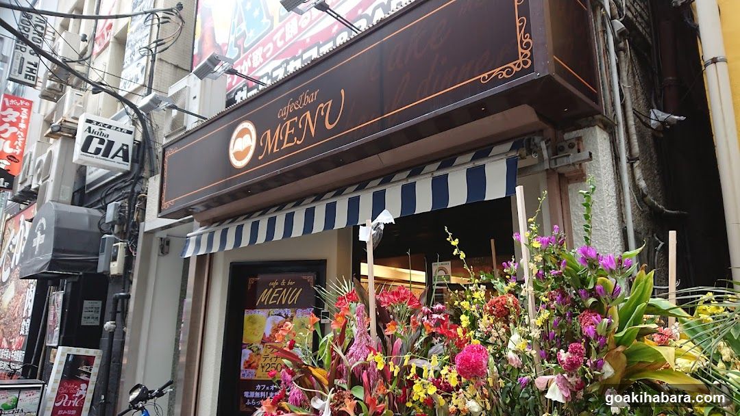 A street-level view of a cafe and bar storefront featuring a dark brown awning with gold lettering reading 'cafe&bar MENU' and decorative borders. Below, a striped blue-and-white canopy extends over the entrance. A large floral arrangement in vibrant colors (pink, purple, yellow) sits at the base of the building. To the left, adjacent signs include 'Media DIA' and other vertical banners. The shop window displays a poster with Japanese text and images of food items, including what appears to be sushi or sashimi. Overhead wires crisscross above the scene.