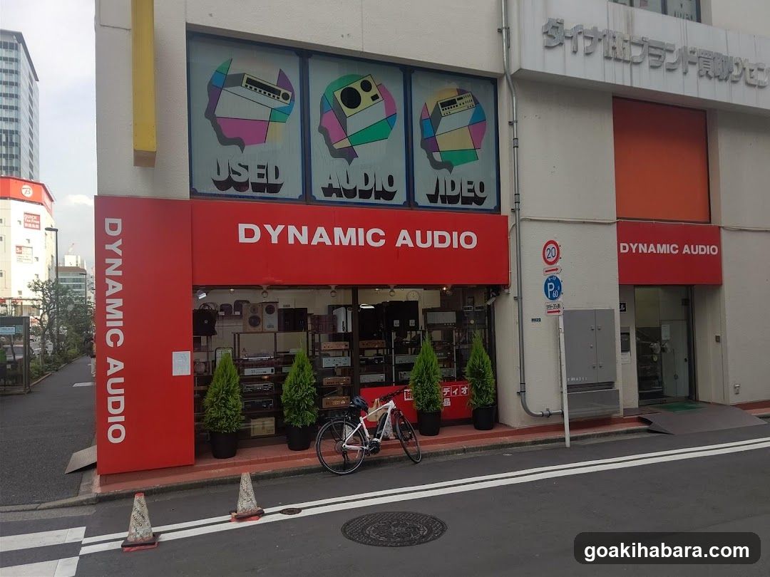 A street-level view of the Dynamic Audio shop, featuring a red facade with large signage reading 'DYNAMIC AUDIO' and 'USED AUDIO VIDEO'. The entrance is flanked by potted plants and a bicycle parked outside. Above the store, there are three windows displaying colorful graphics related to audio equipment.