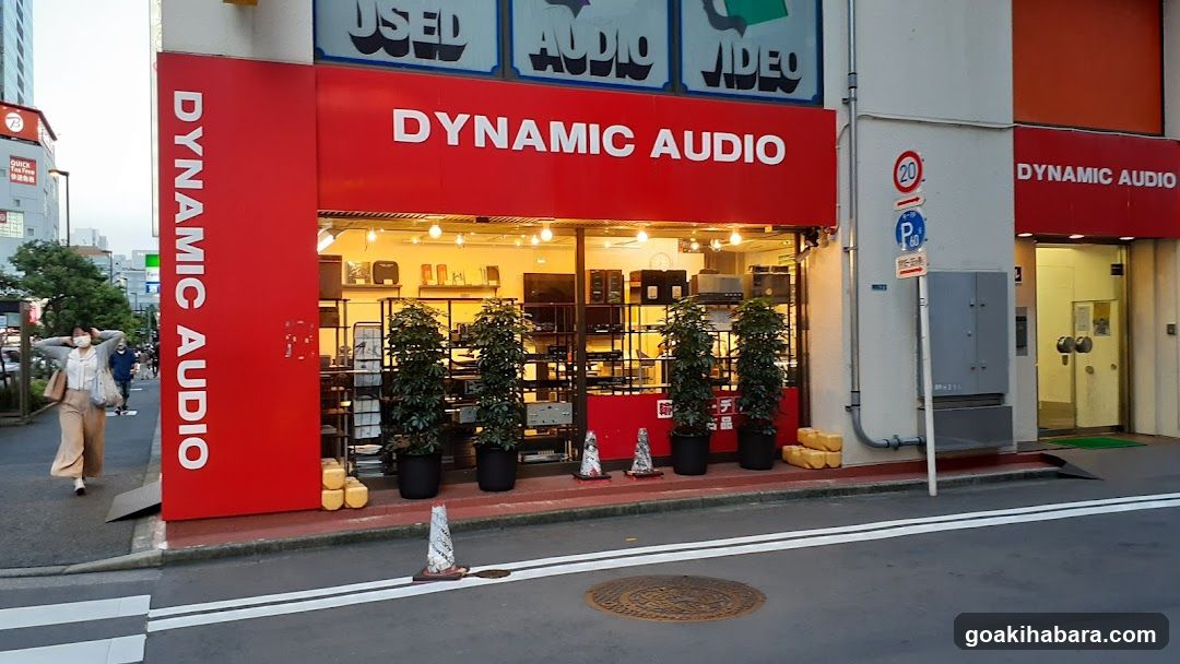 A street-level view of a shop named 'DYNAMIC AUDIO' with prominent red signage displaying the name vertically and horizontally. The storefront features large glass windows revealing shelves filled with audio equipment, including speakers and electronic devices. Potted plants line the entrance, and traffic cones are placed near the sidewalk. A pedestrian walks by on the left side of the frame.