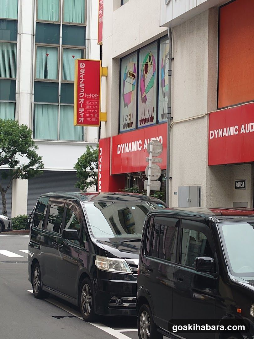 A street-level view of a shop named 'DYNAMIC AUDIO' with red signage and large windows displaying colorful graphics. Two black vans are parked in front.