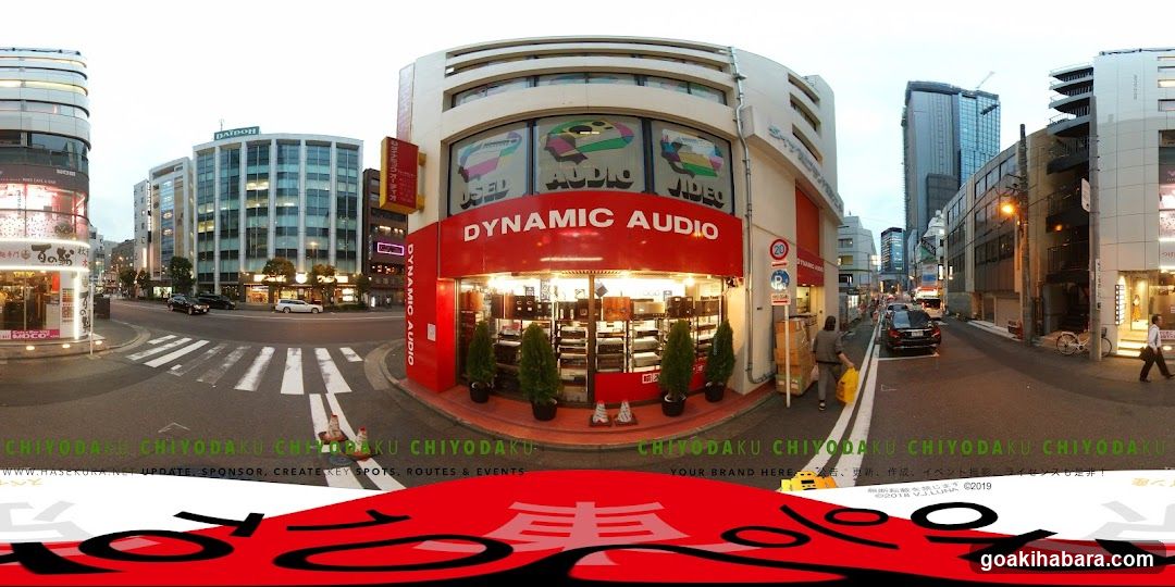 A fisheye view of a street corner in Chiyoda, Tokyo featuring the 'DYNAMIC AUDIO' shop with visible signage and illuminated display windows showing audio equipment. The building is white with red accents, surrounded by other commercial buildings and urban streetscape.