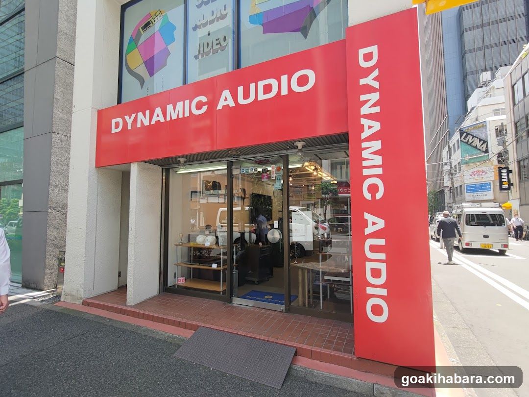 A red storefront with 'DYNAMIC AUDIO' signage on the facade and vertical banner. Glass doors reveal interior shelves, equipment, and a person inside.