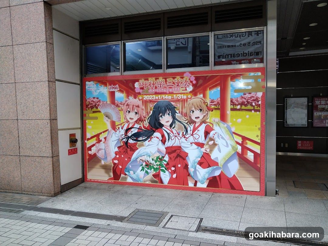 A large promotional poster featuring three anime characters in traditional Japanese attire is displayed on the exterior wall of a building entrance. The poster advertises an event titled '神楽祭' (Kagura Matsuri) scheduled from January 14 to January 31, 2023. Above the main display, smaller signs with Japanese text are visible near the windows.