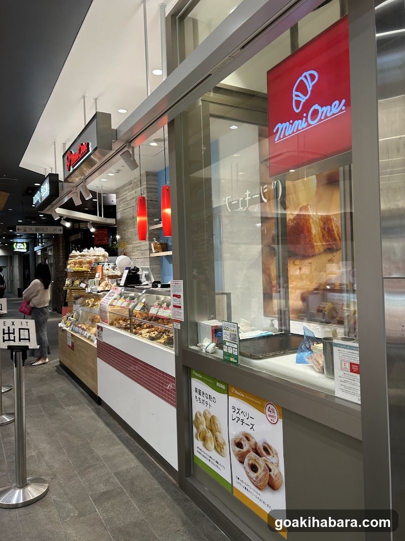 A bakery shop with a glass facade displaying pastries and breads inside. The sign 'Mini One' is visible on the top right of the window. There are posters advertising products like raspberry danishes and mochi buns. A person is walking by in the background near an exit sign.