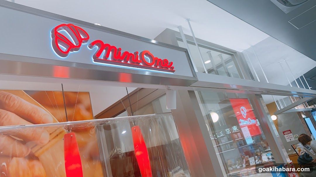 A modern shop front with a prominent red neon sign reading 'Mini One' above the entrance. The storefront features large glass windows displaying promotional posters and interior items. A clear plastic barrier hangs at the entrance, likely for safety or hygiene purposes. Inside, shelves stocked with products are visible through the glass.