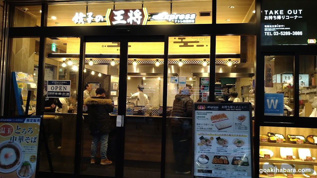 A brightly lit storefront with large glass windows and doors, displaying illuminated signage in Japanese and English. Inside, staff are visible behind the counter, preparing food. A customer stands near the entrance. Outside, a menu board lists various dishes with prices.