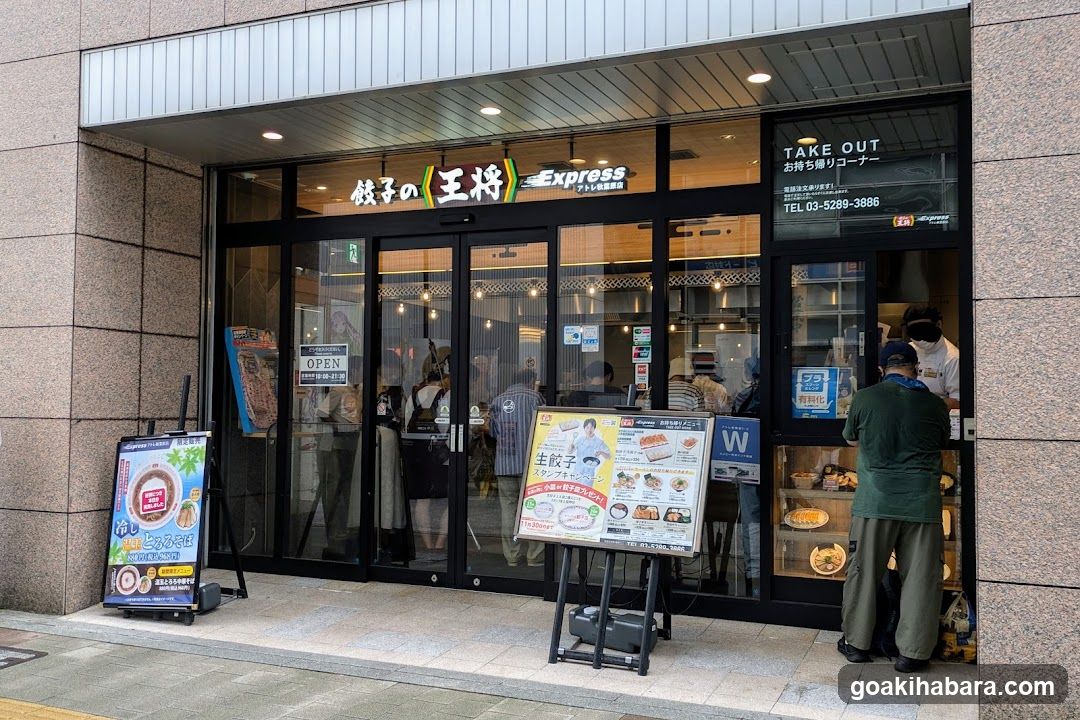 A modern storefront with large glass windows and doors, featuring signage reading 'Noodles of the King Express' in Japanese and English. A customer is ordering at the counter inside, while promotional boards display menu items like dumplings and noodles.