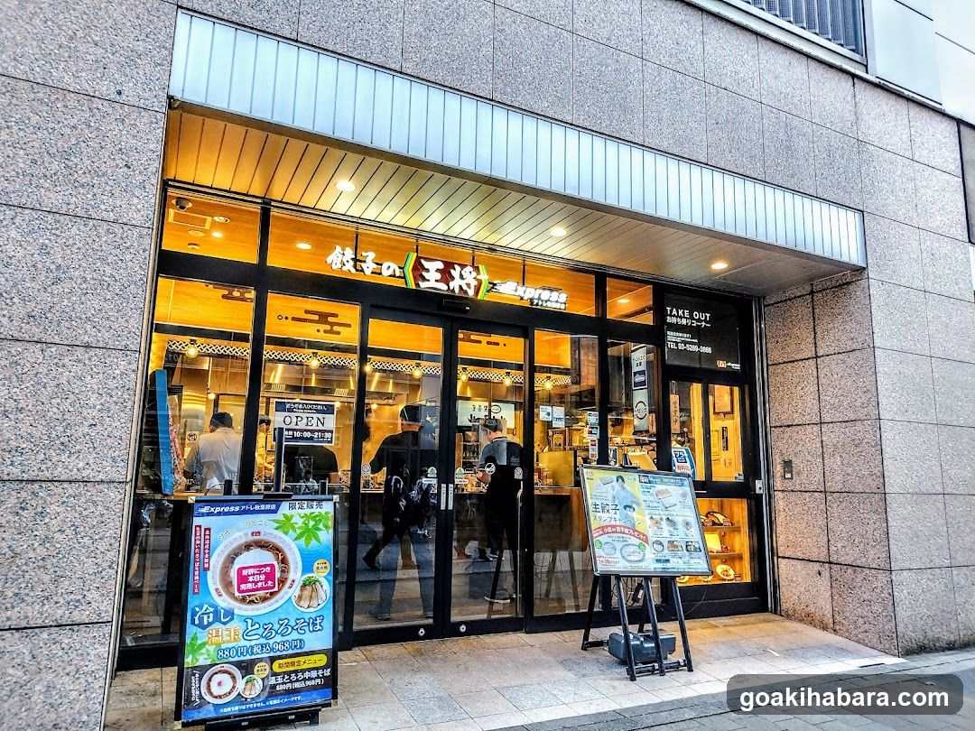 A modern shop front with large glass windows and doors, displaying warm interior lighting. Visible signage includes Japanese characters above the entrance and a 'TAKE OUT' sign on the right side. Two menu boards stand outside: one on the left featuring gyoza promotions and another on the right showing various food items.