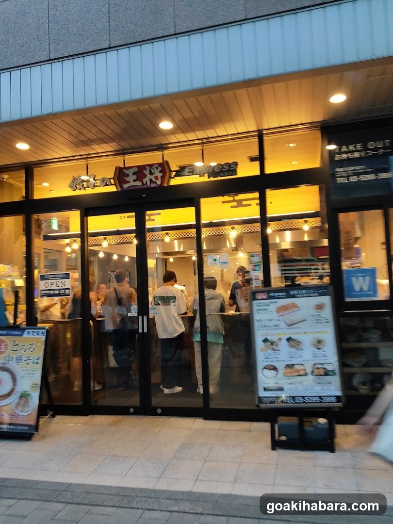 The photo shows the exterior of a restaurant entrance at night, illuminated by warm interior lighting. Several customers are standing in line or waiting inside through glass doors. A sign above reads 'WINGRESS' and another displays Japanese characters. To the left, an open sign is visible on the door. On the right side, there's a menu board displaying various food items with prices.