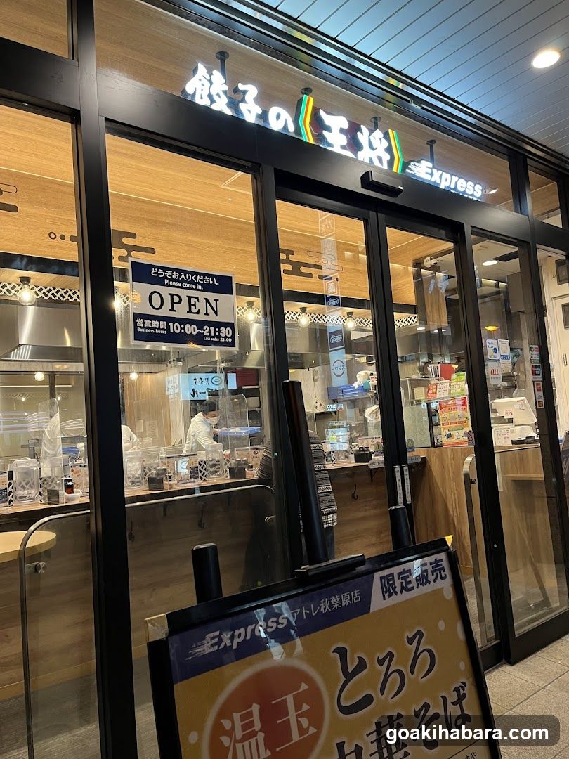 A storefront with glass doors and windows displaying the illuminated sign '餃子の王将 Express'. Inside, staff are visible behind a counter. A sign indicates opening hours from 10:00 to 21:30. Outside, there is a promotional board advertising limited-time items like 'Wagyu Soba' and 'Hot Tamago'.