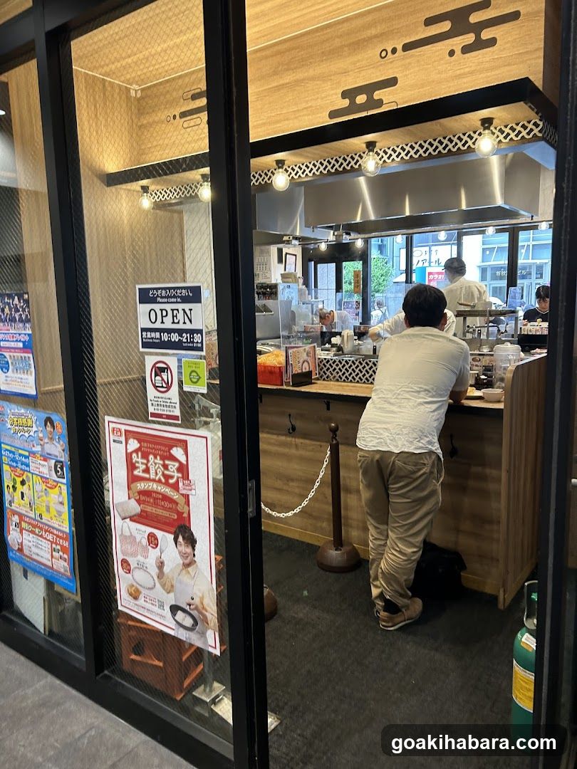 A photo of a restaurant's exterior entrance, showing glass doors and windows. Visible signage includes an 'OPEN' sign indicating operating hours from 10:00 to 21:30 on weekdays. Inside, customers are seen ordering at the counter, and staff members are preparing food in the kitchen area. Posters and advertisements are displayed on the glass doors.