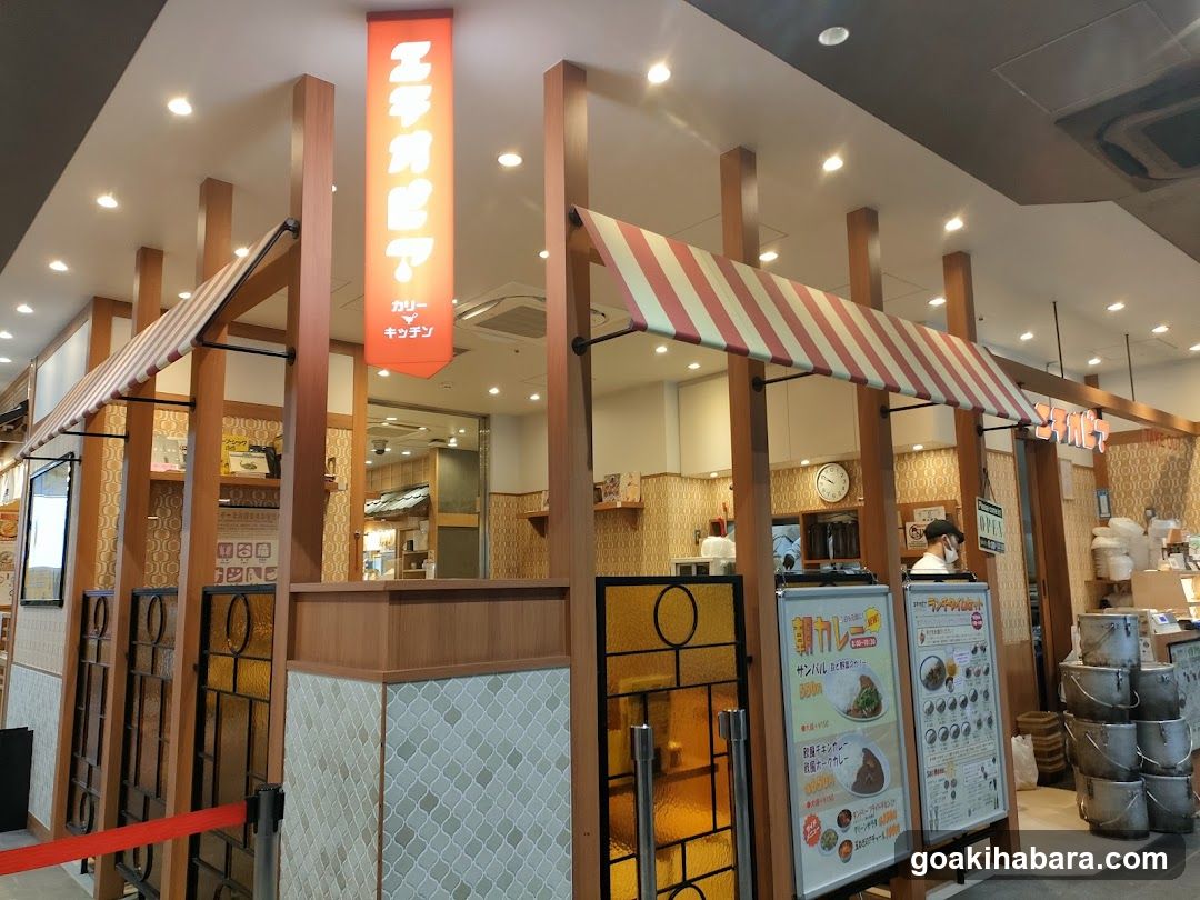 The image shows the storefront of a ramen shop with wooden pillars and striped awnings. A vertical orange sign displays Japanese characters, likely indicating the restaurant name. Inside, there are tables and chairs visible through glass windows. Outside, there are several metal buckets stacked near the entrance. Two menu boards stand outside the shop, displaying various dishes and prices.