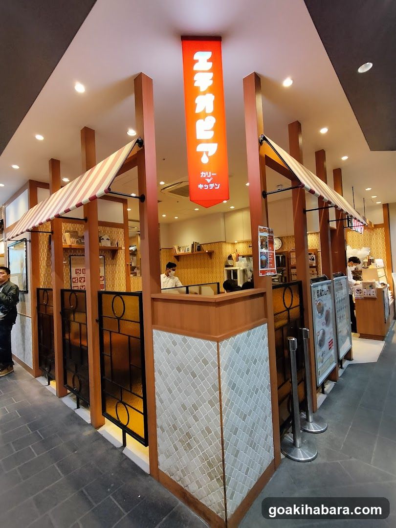 The image shows the exterior of a shop with a vertical orange sign displaying Japanese characters and 'Curry' in English. The storefront features striped awnings, wooden pillars, and tiled walls. Inside, staff are visible behind counters, and menu boards stand outside.