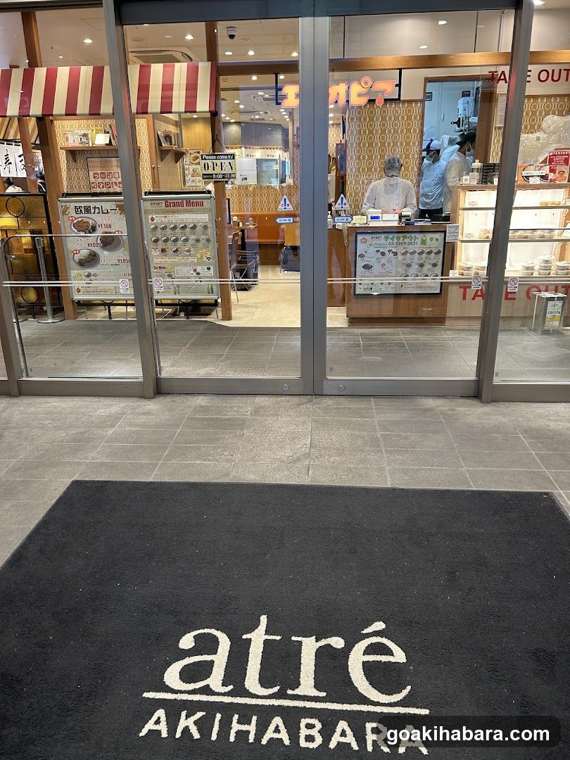 Photo of the physical exterior entrance to a shop, featuring glass doors with visible signage including 'OPEN' and menu boards inside. The floor mat reads 'atré AKIHABARA'. Staff are visible behind the counter.