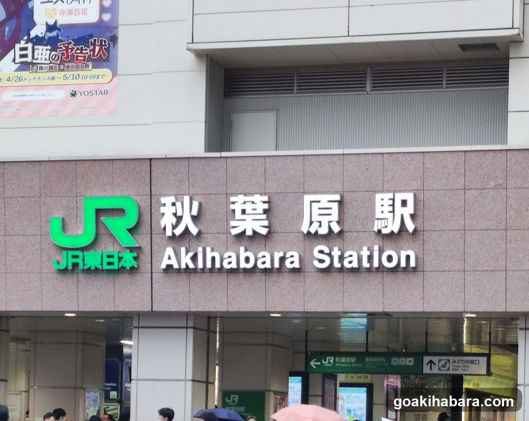 Photo of the exterior entrance facade of Akihabara Station, featuring prominent signage with 'JR' logo and Japanese/English station name. Visible posters above include promotional material for an event on April 26th.