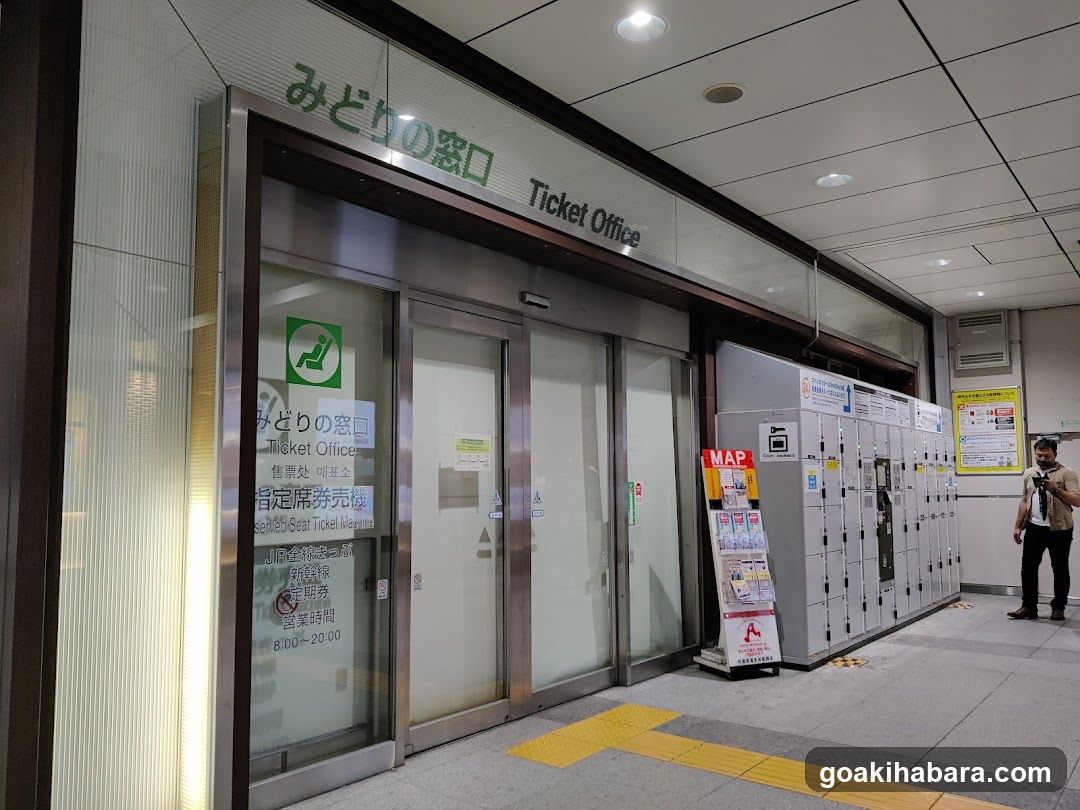 A modern ticket office entrance with glass doors and signage reading 'Midori no Kuchi' in Japanese and English. Adjacent to the entrance are several lockers and a promotional stand displaying maps and information.