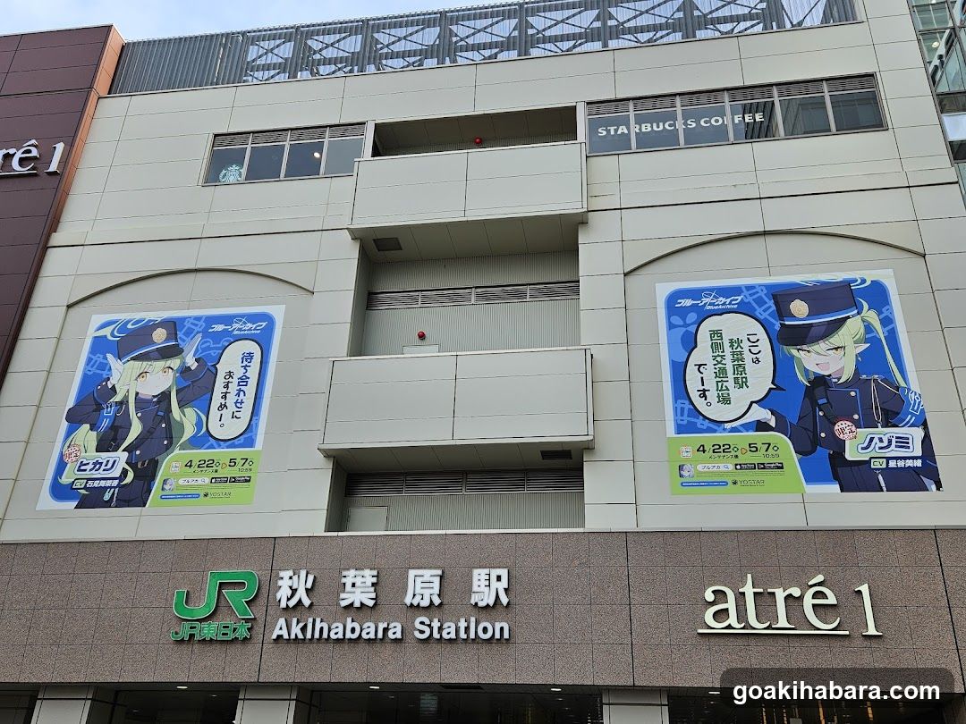 Exterior facade of Akihabara Station featuring a multi-story building with visible signage including 'JR East Japan', '秋葉原駅' (Akihabara Station), and 'atré 1'. Two large posters advertise anime characters in police uniforms, promoting an event from April 22 to May 7. The Starbucks Coffee sign is also visible on the upper level.