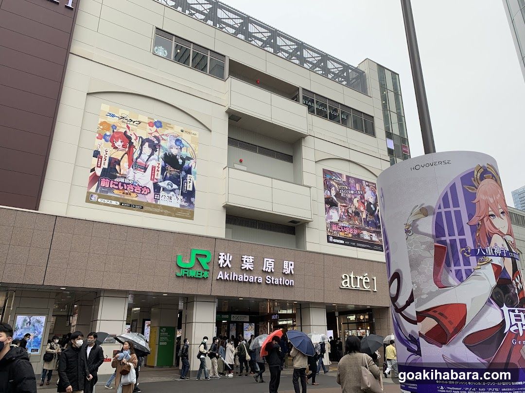 Photo of the exterior entrance to Akihabara Station, featuring JR and atré1 signage on the building facade. Large anime-themed posters are displayed above the entrance, including one for 'Genshin Impact' and another with multiple characters. People are walking in front of the station.